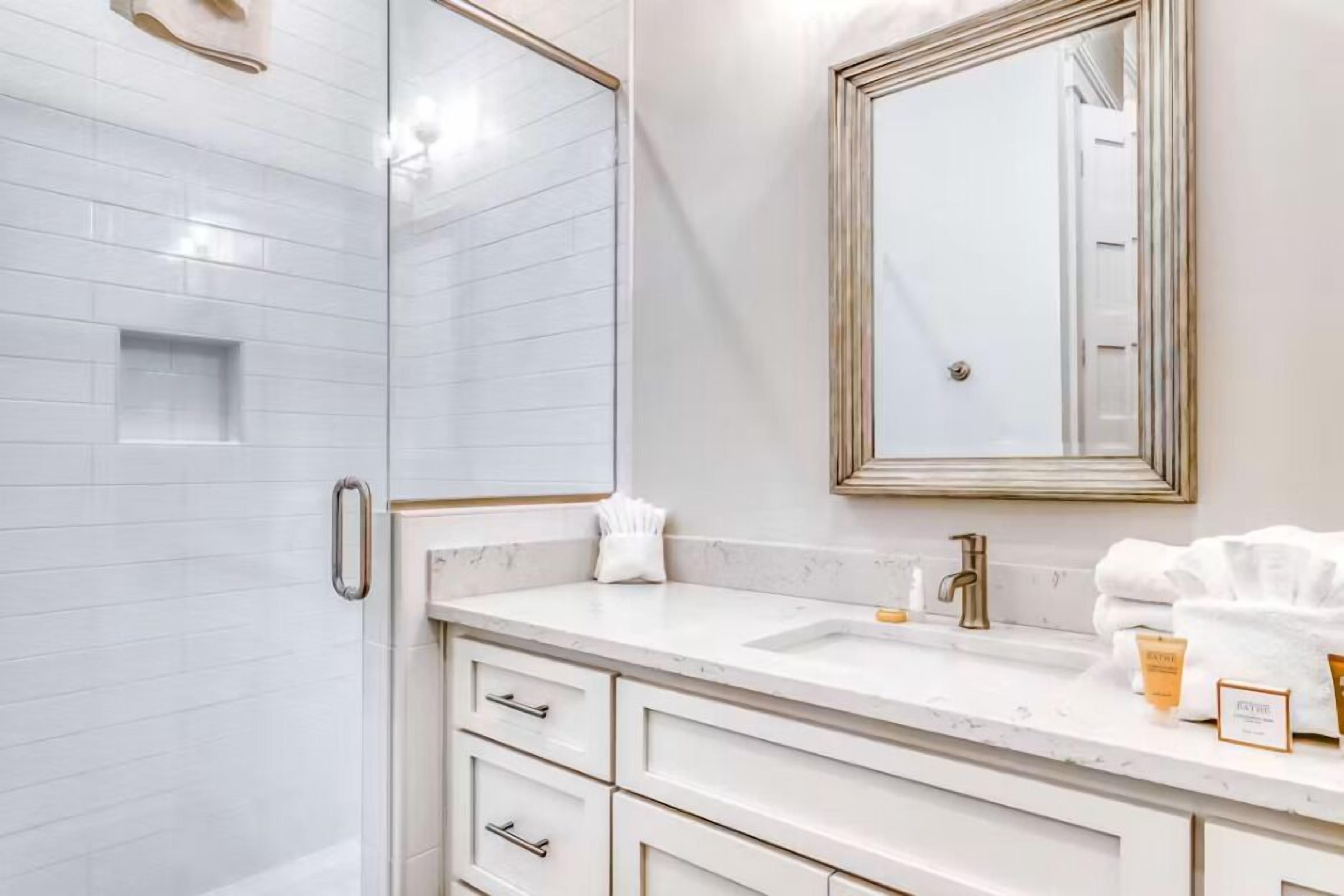 A bathroom with a white tiled shower, a vanity with a mirror, and a sink with toiletries.