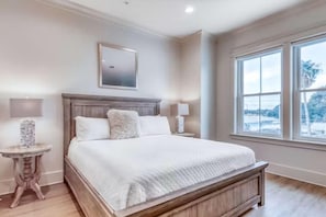 Une chambre avec un grand lit, deux tables de chevet, une commode, un tableau encadré au mur et une fenêtre donnant sur un beau paysage.