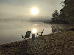 Rauhallinen maisema järven rannalla, jossa on kaksi Adirondack-tyylistä tuolia järvelle päin, ja josta avautuu selkeä näkymä auringon heijastukseen järven pinnalla sekä vehreä rantaviiva.