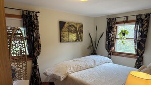A bedroom with a bed, a window with curtains, a potted plant, and a wall-mounted artwork.