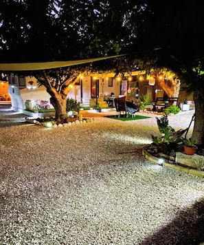 Un espace extérieur aménagé pour la nuit, avec un chemin de gravier, des arbres et un bâtiment doté d’une terrasse.