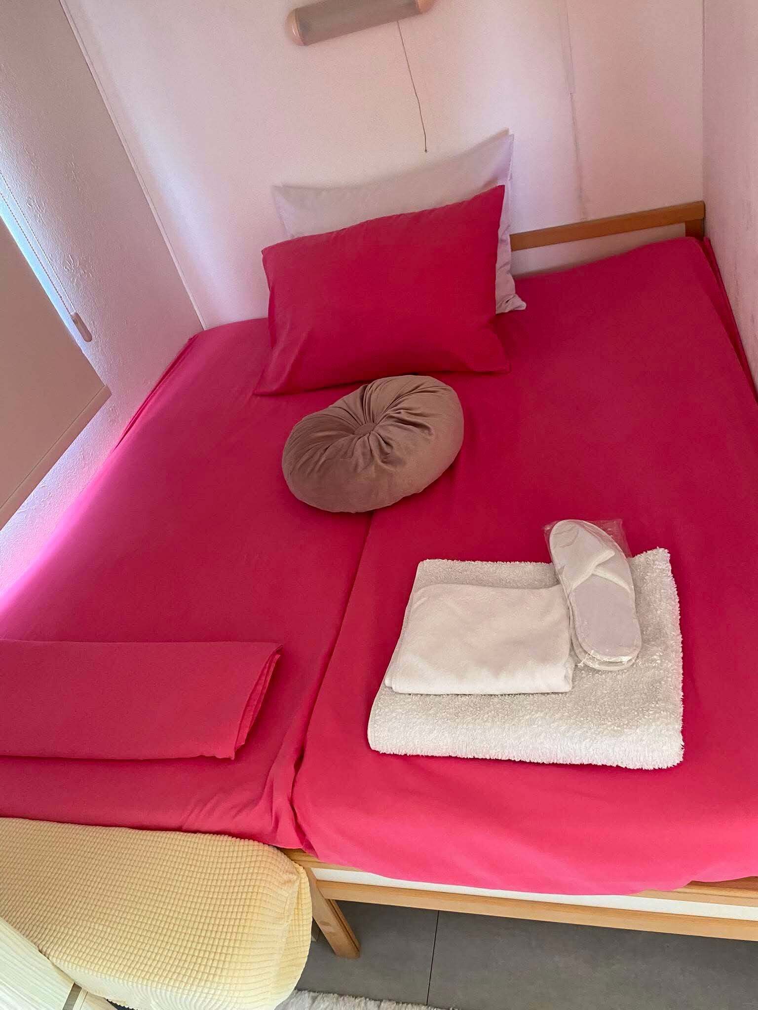 Una cama individual con una colcha roja, una almohada color beige y una toalla blanca, con un cabecero de madera.