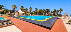 En swimmingpool med fliseterrasse, palmer og strandparasoller.