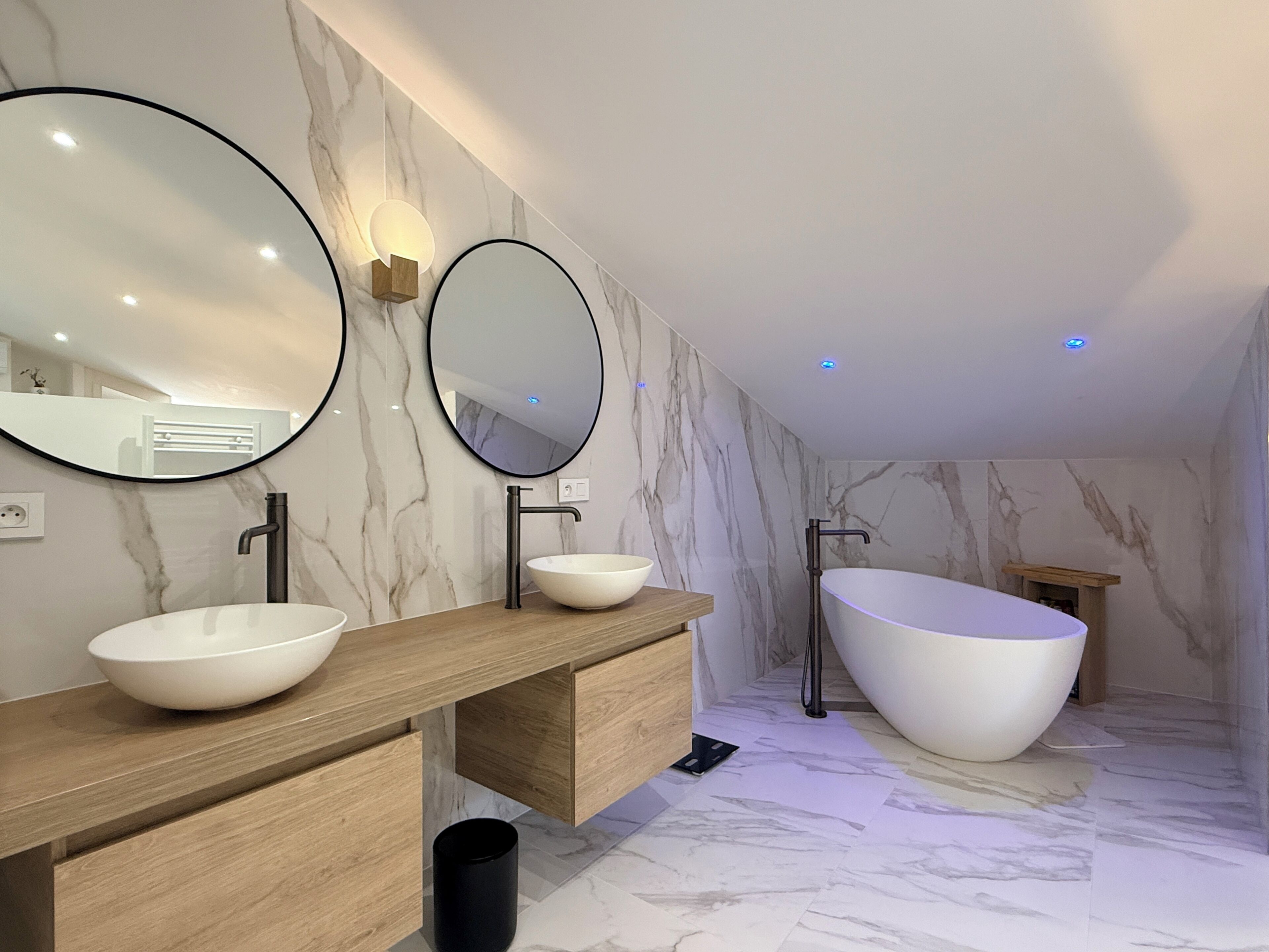 A modern bathroom with a freestanding bathtub, two sinks with round mirrors, and marble walls.