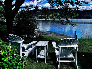 Eine Uferlandschaft mit Adirondack-Stühlen und einem Picknicktisch, umgeben von viel Grün und Blick auf den See.