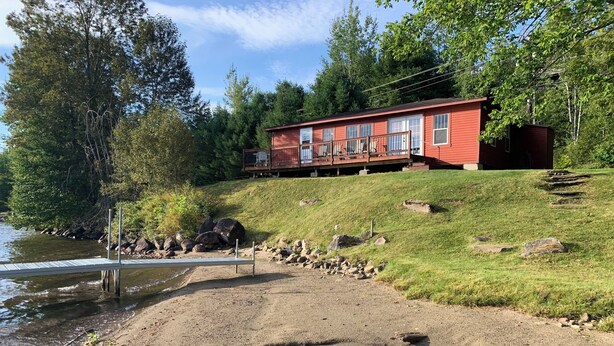 En röd stuga med en veranda, belägen på en gräsklädd kulle vid en sjö.