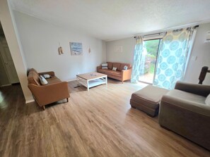 Een woonkamer met een bruine bank, een witte salontafel en een bruin voetenbankje. Er is een schuifdeur die uitkomt op een buitenruimte.