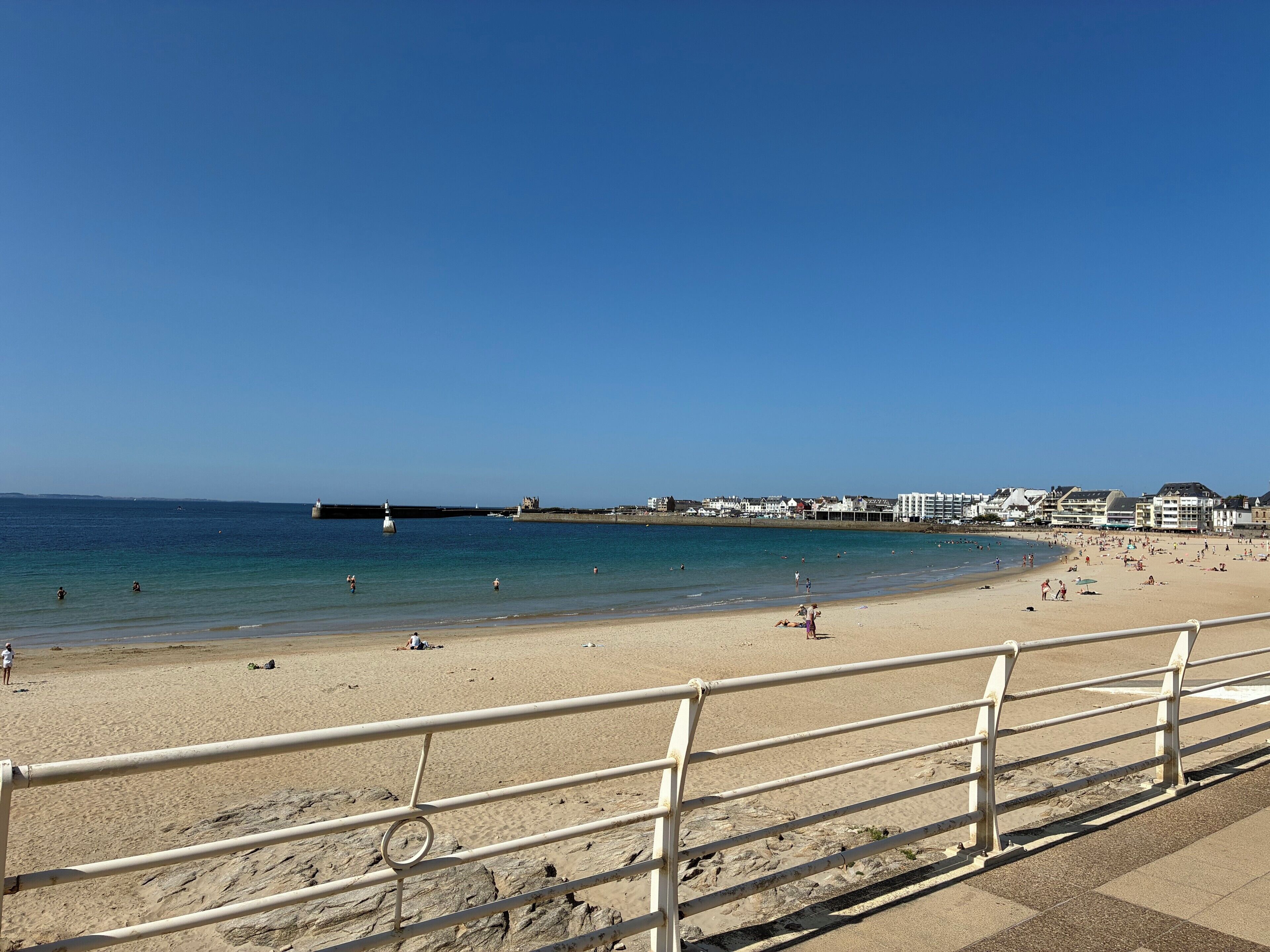 A beach with a clear sky, a sandy shore, and a metal railing in the foreground.
