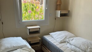 Ein Schlafzimmer mit zwei Betten, einem Fenster mit Blick auf Blumen, einer Lampe auf einem Regal und einer Steckdose.