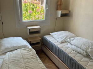 Ein Schlafzimmer mit zwei Betten, einem Fenster mit Blick auf Blumen, einer Lampe auf einem Regal und einer Steckdose.