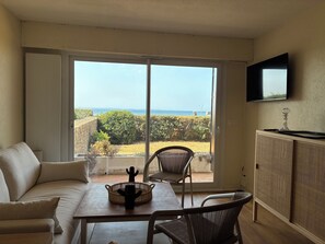 A room with a large sliding glass door leading to a balcony with a view of the sea. There is a sofa, a dining table with chairs, and a cabinet with a TV mounted above it. - Appartement plage et vue mer (Quiberon)