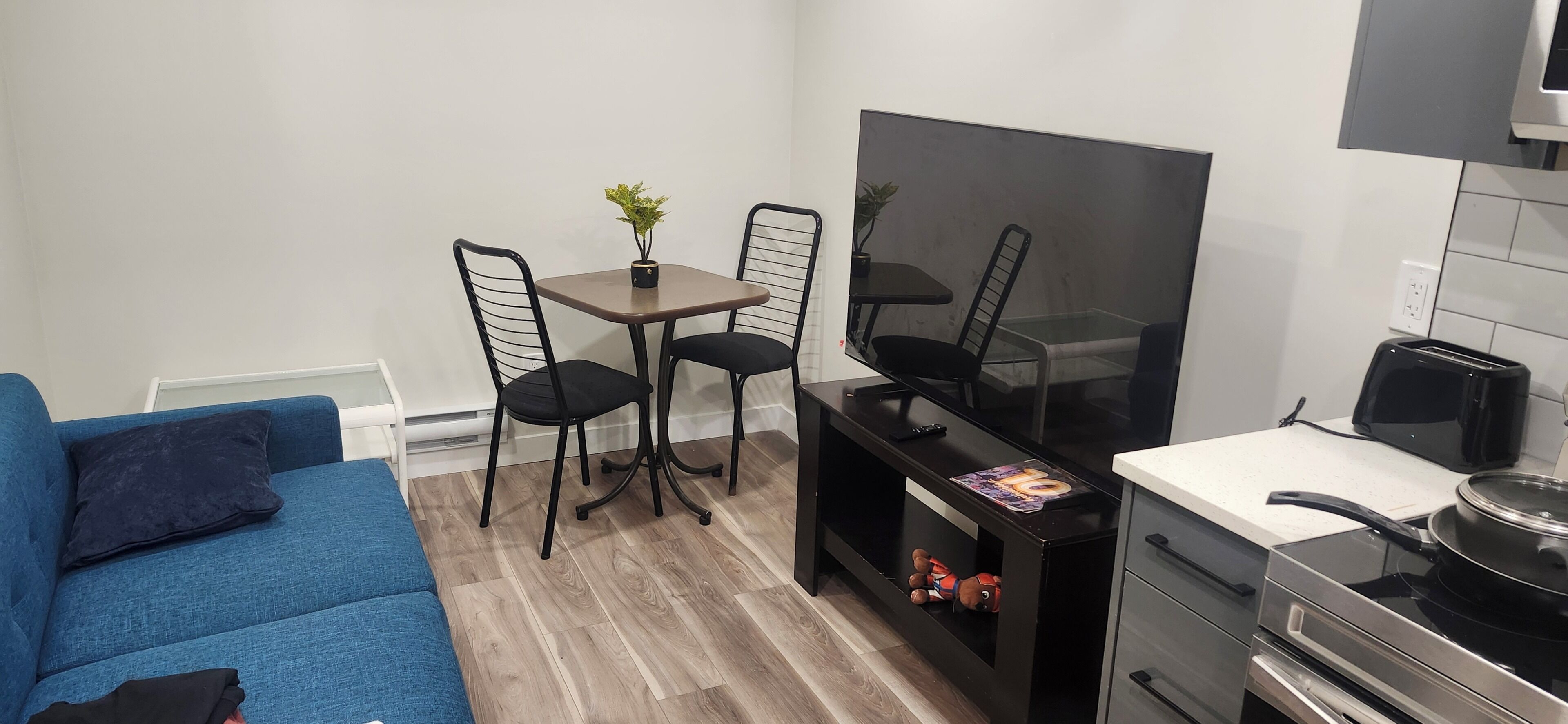 Un espacio habitable compacto con un sofá azul, una pequeña mesa de comedor con dos sillas, un televisor de pantalla plana, un tostador y una estufa.