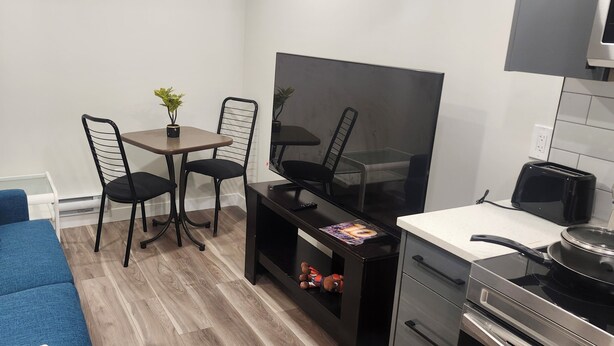 Een compacte leefruimte met een blauwe bank, een eettafel met twee stoelen, een flatscreen televisie, een broodrooster en een fornuis.