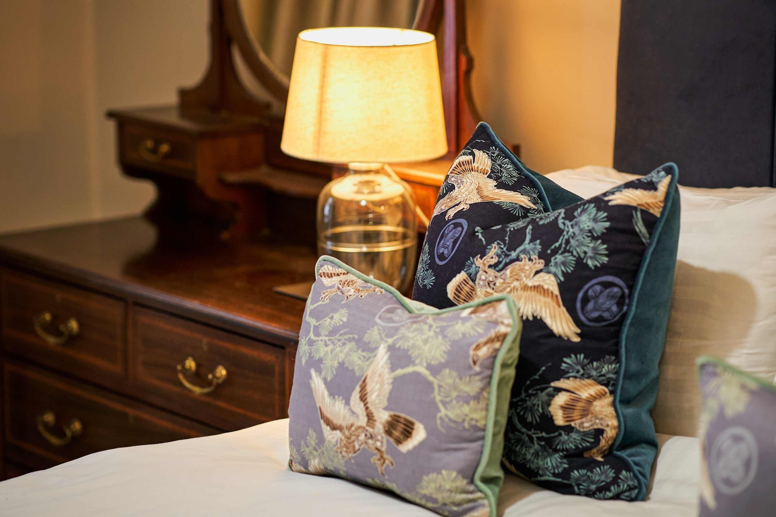 A bed with decorative pillows featuring bird designs, a bedside lamp, and a wooden nightstand with drawers.