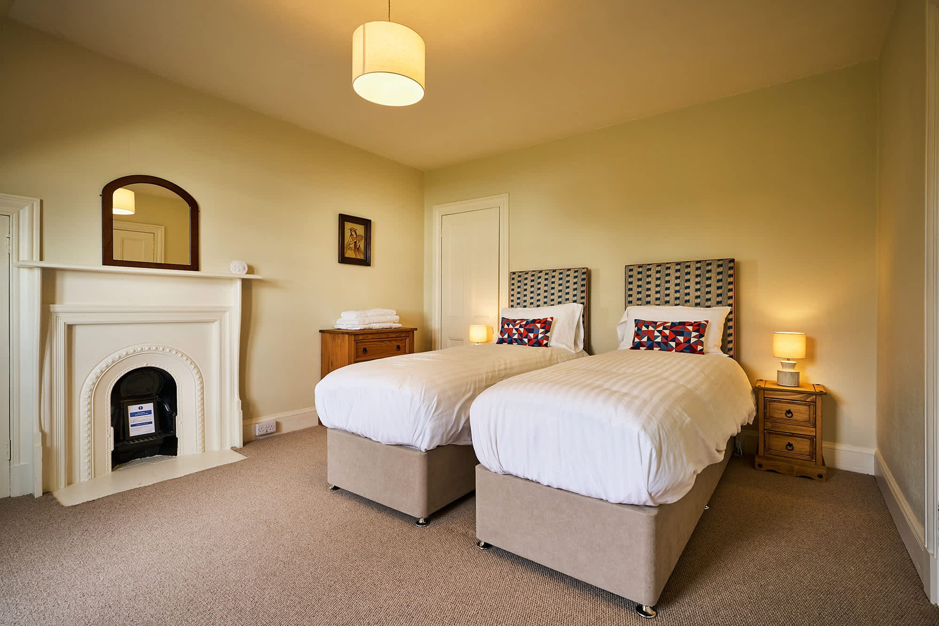 A hotel room with two beds, a fireplace, a nightstand with a lamp, and a mirror.