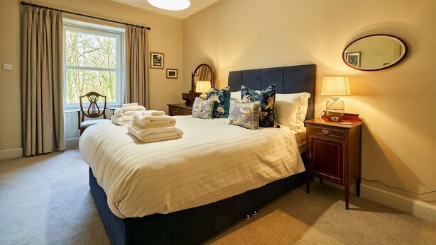 A neatly made bed with a headboard, a bedside table with a lamp, a mirror, and a window with curtains. - Kinclune House Buildings on Private Angus Estate (Kirriemuir)
