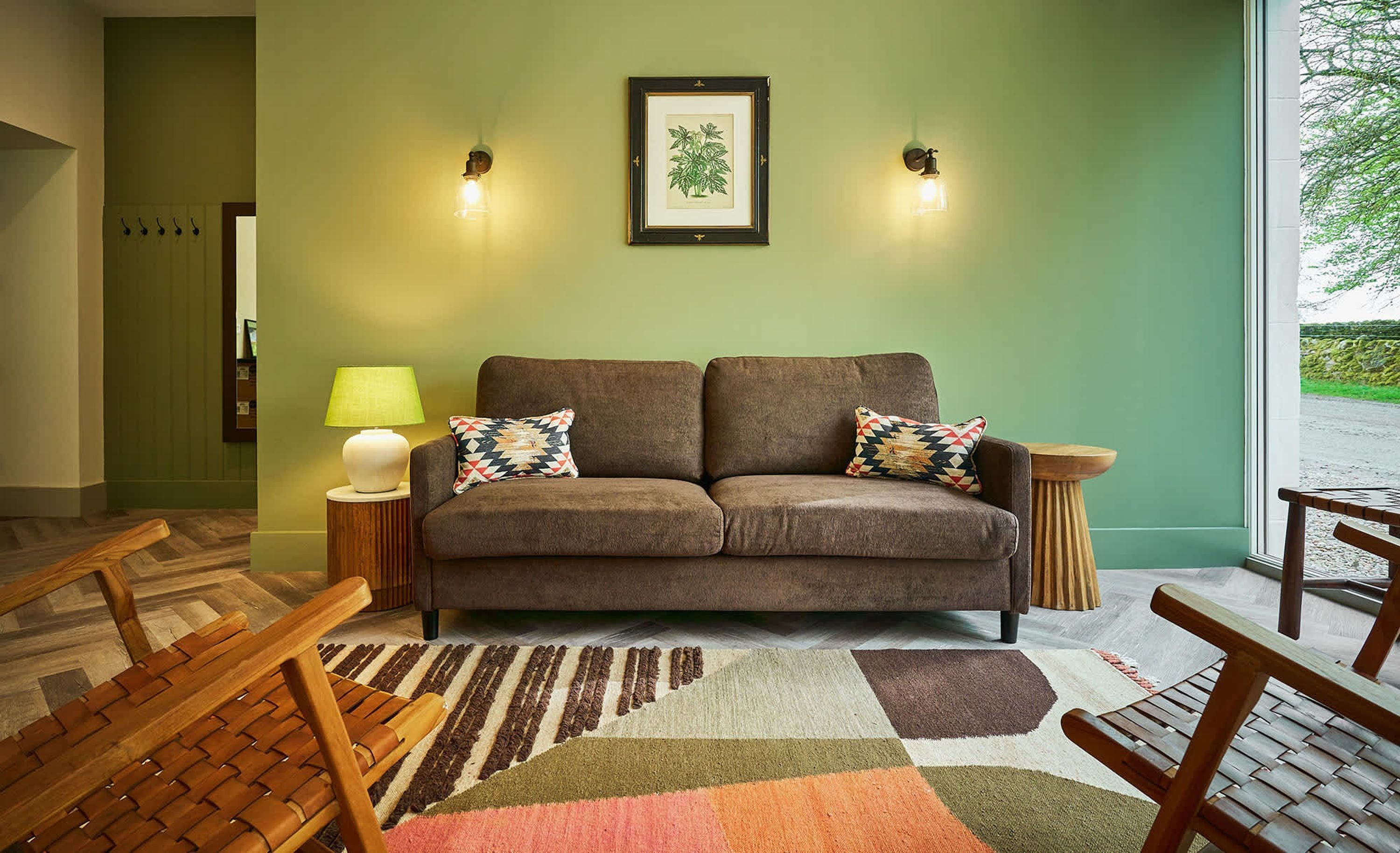 A living room with a brown sofa, two patterned throw pillows, a green wall, a framed botanical print, and a lamp on a side table.