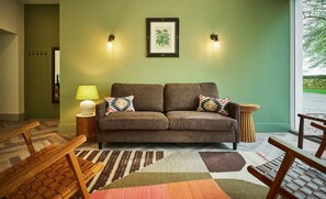 Un salon avec un canapé marron, deux coussins décoratifs à motifs, un mur vert, une estampe botanique encadrée et une lampe posée sur une table d’appoint.