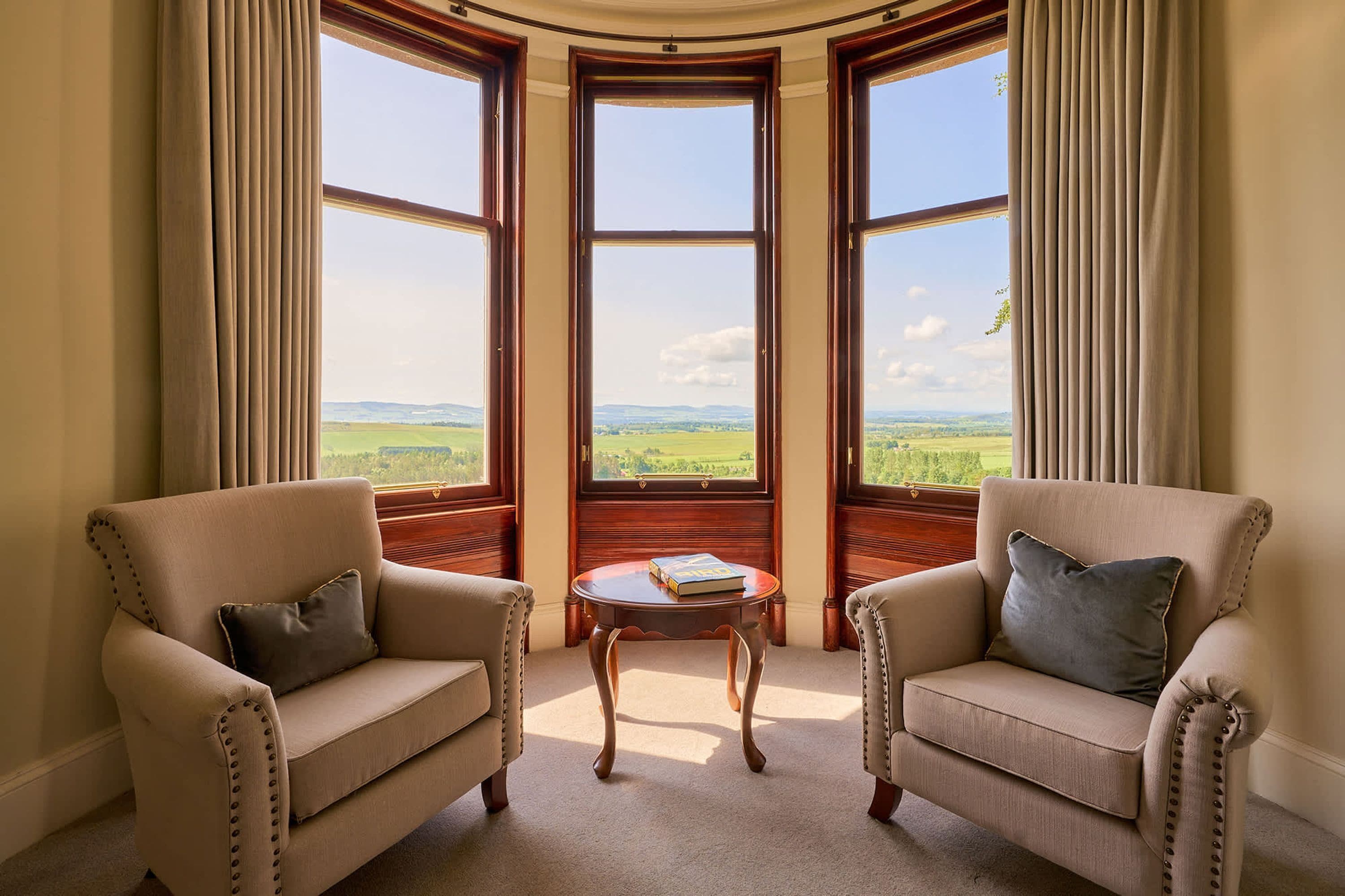 A room with two armchairs, a small table, and a bay window with a view of the countryside.