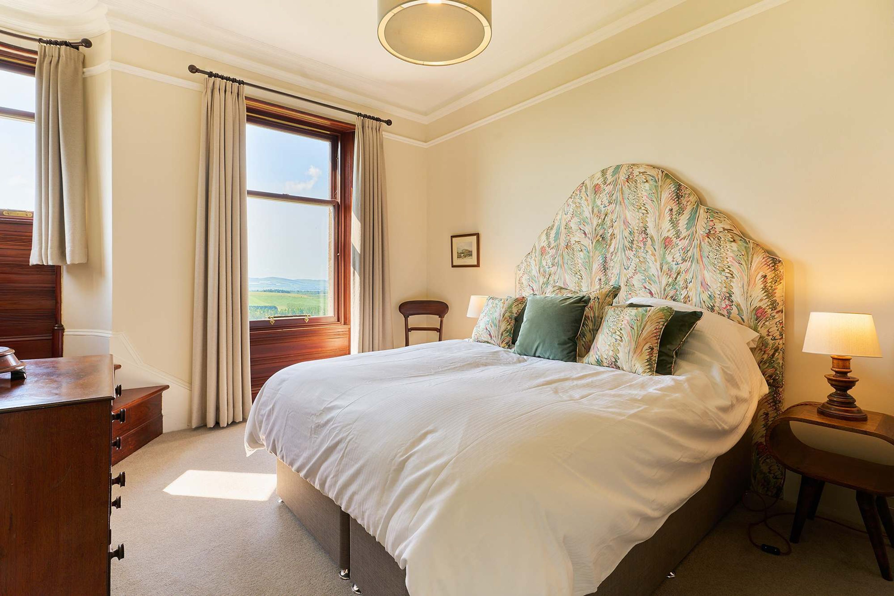 A bedroom with a large bed, a wooden dresser, a nightstand with a lamp, and a window with curtains.