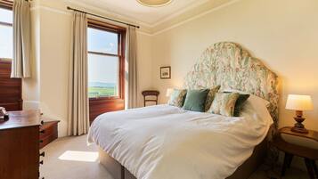 A bedroom with a large bed, a wooden dresser, a nightstand with a lamp, and a window with curtains.