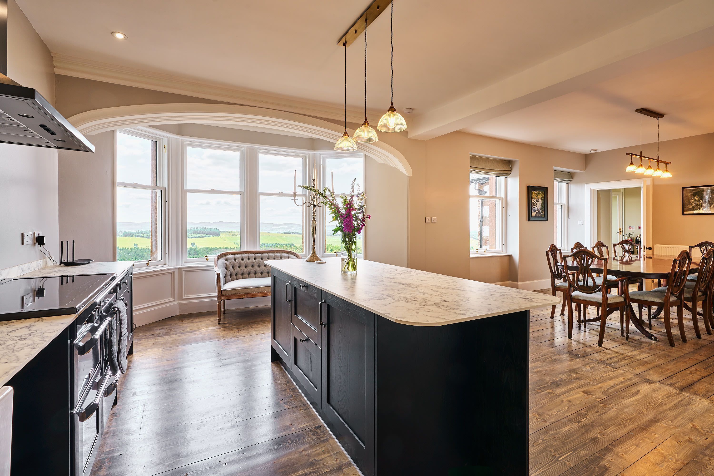 A modern kitchen with a large island, wooden flooring, and a view of the countryside through large windows.
