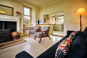 A living room with a fireplace, two armchairs, a sofa, and a kitchen area visible through an open door. - Peaceful Farmhouse Kinclune Estate Angus Glens (Kirriemuir)