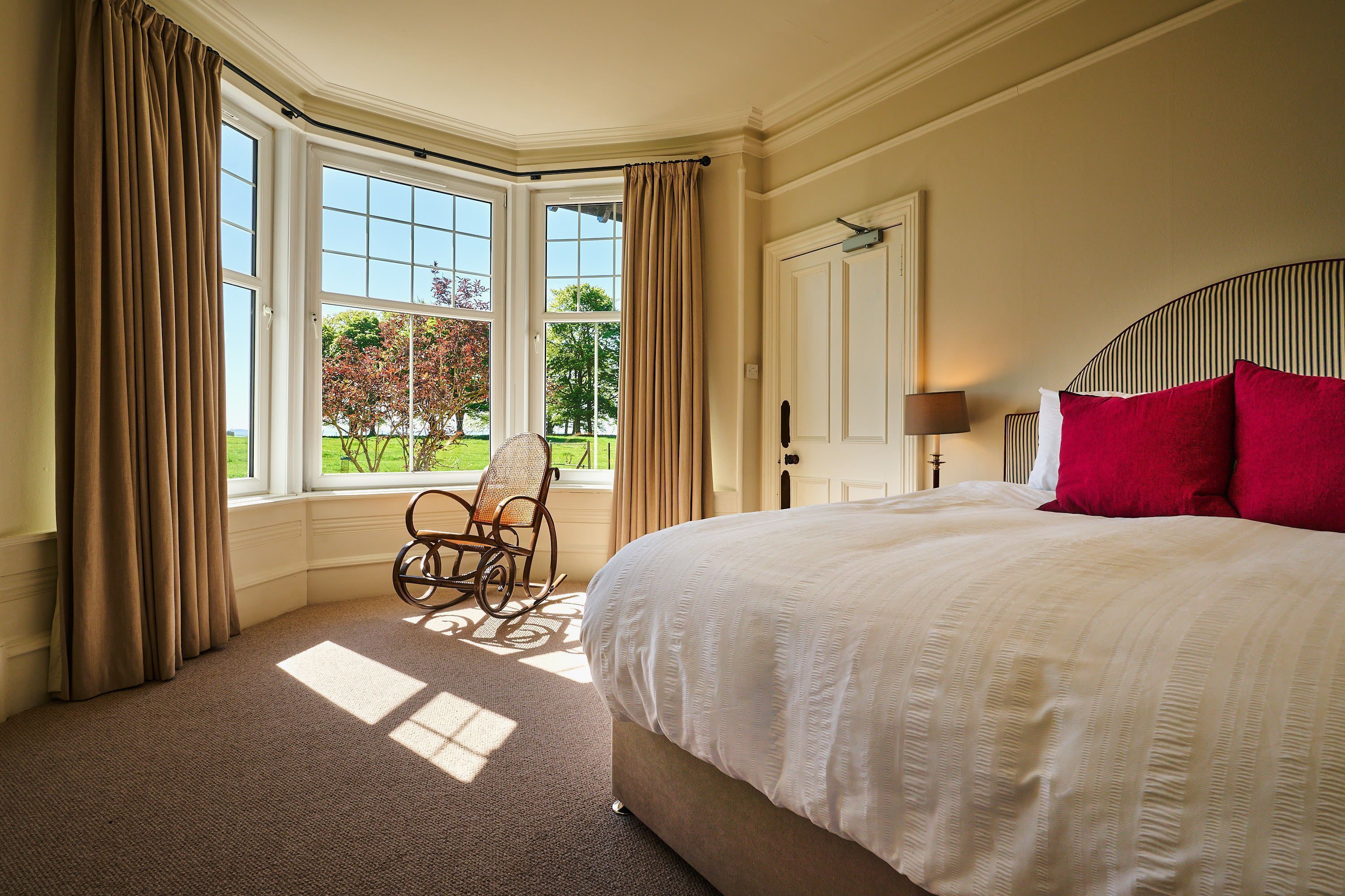 A bedroom with a large bed, a chair, a window with curtains, and a view of trees.