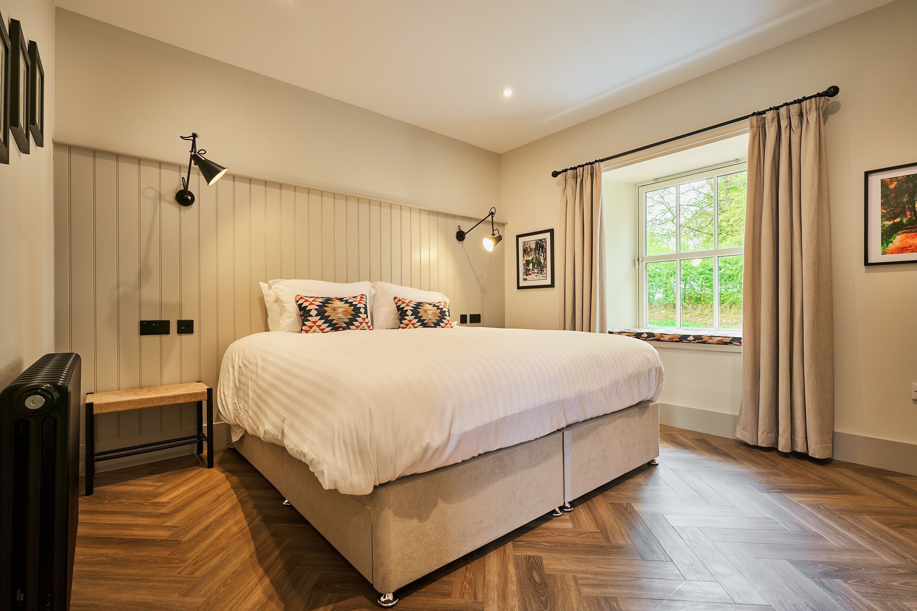 Une chambre à coucher avec un grand lit, deux lampes de chevet, un banc, un radiateur et une fenêtre avec des rideaux.