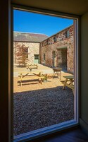 Vista a través de una ventana que muestra un patio con edificios de piedra, mesas de picnic y suelo de grava.