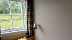 A single bed with a white mattress and a yellow pillow, a window with a view of greenery, and a wall-mounted lamp. - Apartment in Stamsund (Stamsund)