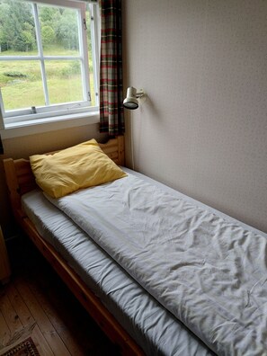 A single bed with a white mattress and a yellow pillow, a window with a view of greenery, and a wall-mounted lamp. - Apartment in Stamsund (Stamsund)