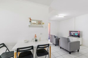 A modern living room with a white dining table, two black chairs, a grey sofa, and a television on a white cabinet.