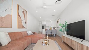 A modern living room with a sofa, a coffee table, a TV, and a dining area in the background. - Brand-New Premium Retreat|Pool &Gym| Parking|Train (Mitchelton)