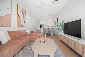 A modern living room with a sofa, a coffee table, a TV, and a dining area in the background. - Brand-New Premium Retreat|Pool &Gym| Parking|Train (Mitchelton)