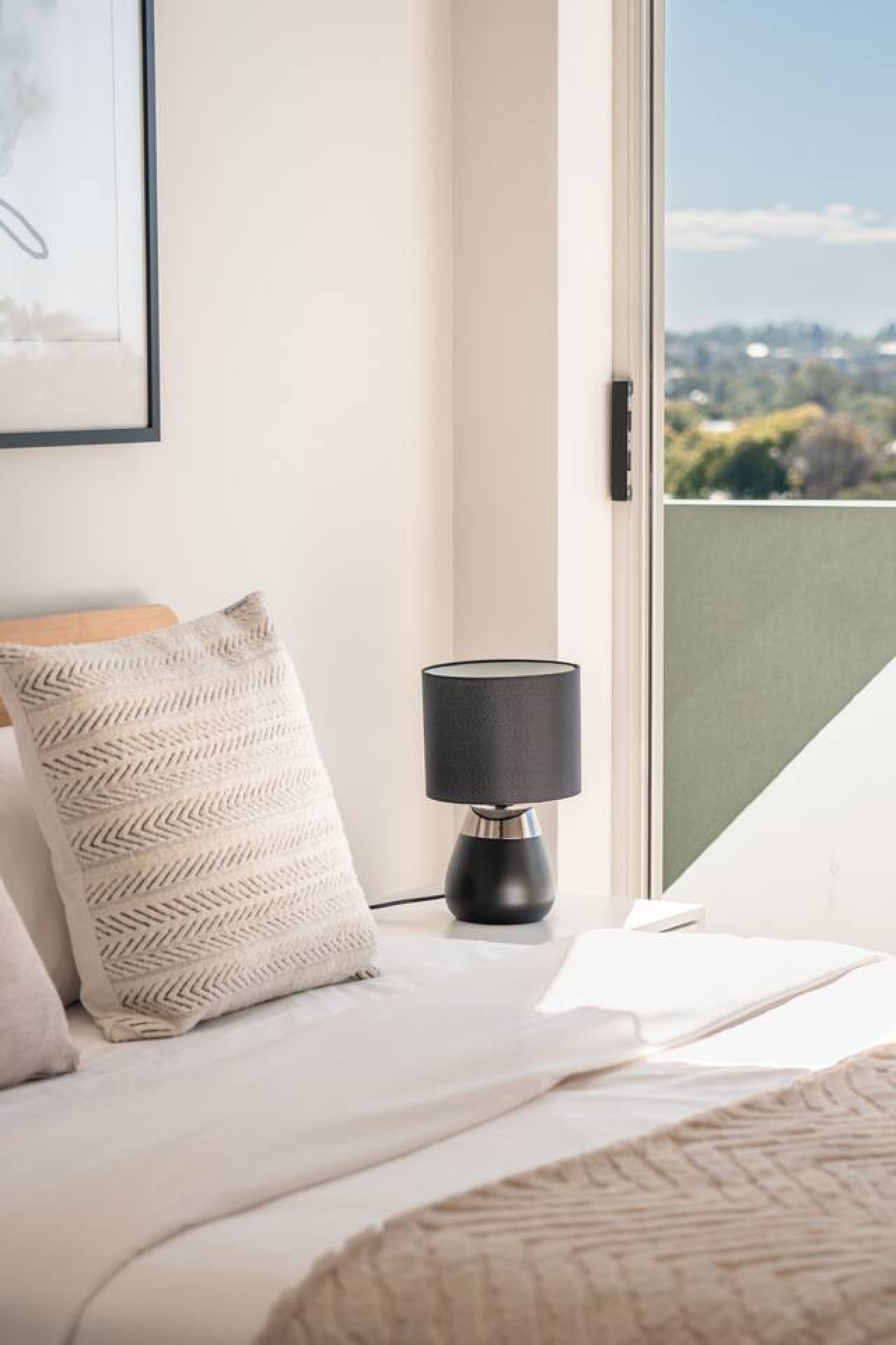 A bedroom with a bed, a pillow, a lamp, and a window with a view of greenery.