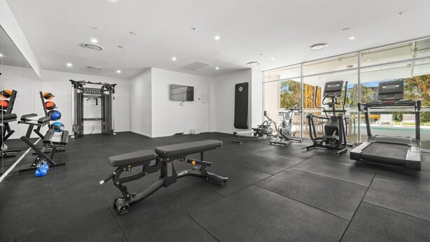 A gym with various exercise equipment, a bench, and a large window with a view. - Brand-New Premium Retreat|Pool &Gym| Parking|Train (Mitchelton)