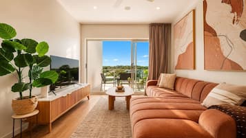 A modern living room with a sofa, a coffee table, and a TV. There is a large window with a view of the outdoors.