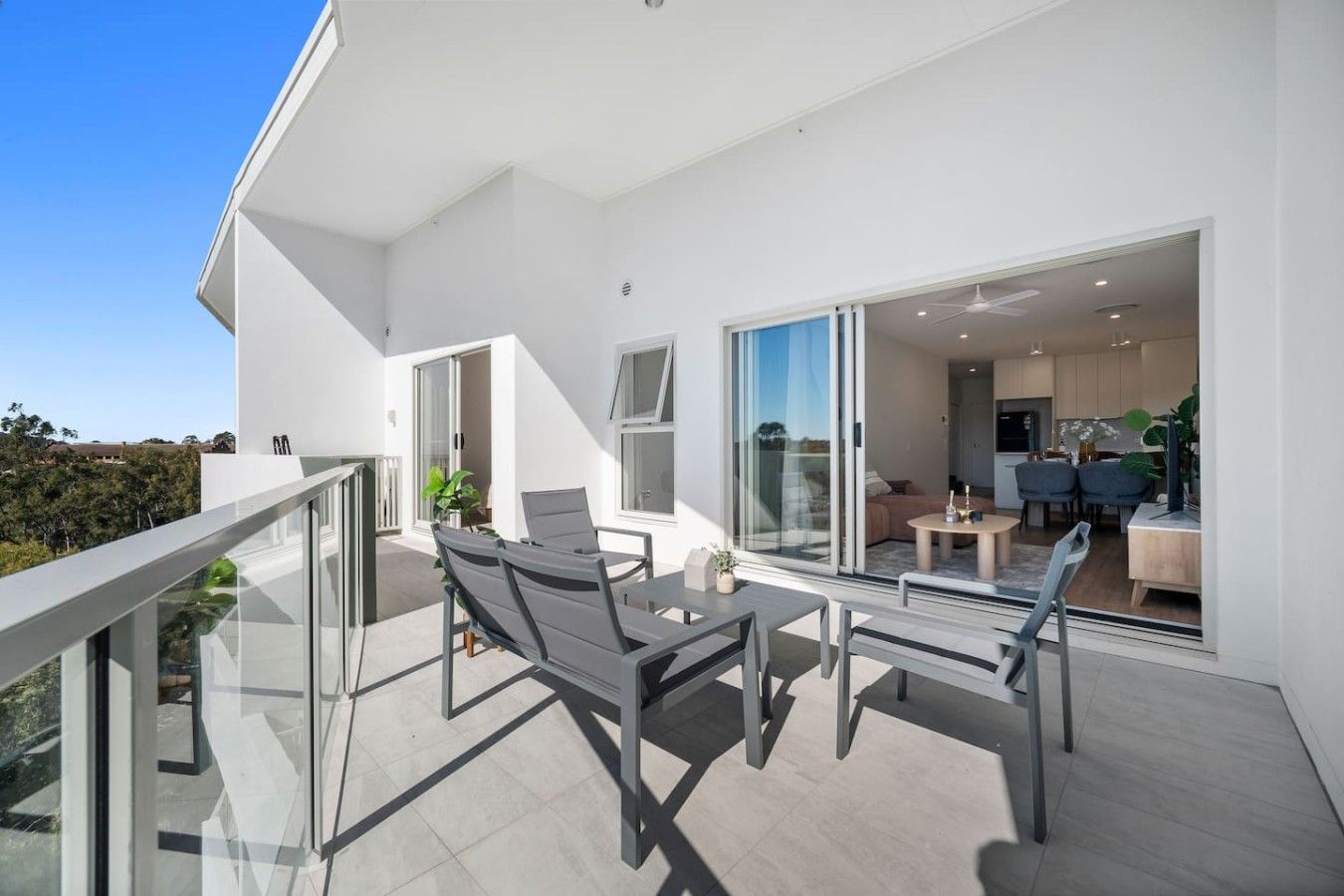 A modern outdoor balcony with a glass railing, a table with chairs, and a view of the interior.
