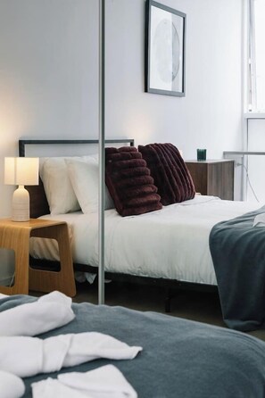 Een hotelkamer met een bed, een nachtkastje met een lamp, een stoel, een groot raam en een lijst met een afbeelding aan de muur.