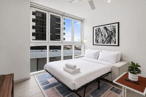 A modern hotel room with a large window, a bed with white linens, a bedside table with a plant, and a framed abstract artwork on the wall. - The Valley Great Value| Sofa Bed |Parking|Pool&Gym (Fortitude Valley)