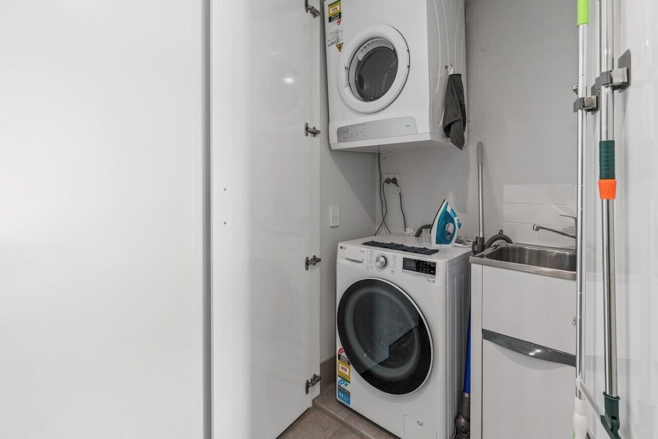 A compact laundry room with a front-loading washer and dryer, a small sink, and a green-handled ironing board.