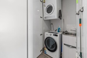 Een praktische wasruimte met een voorlader wasmachine en droger, een klein wastafeltje en een strijkplank met een groene handgreep.