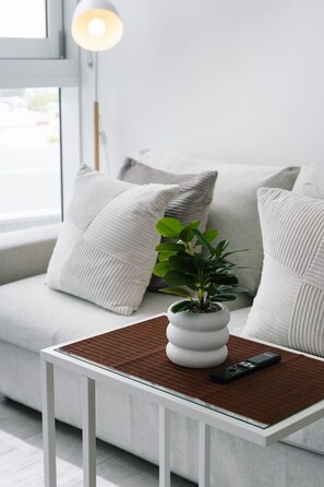 En moderne stue med en hvid sofa, et lille hvidt bord, en potteplante og en fjernbetjening.