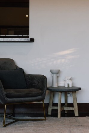 A dark-colored armchair with a gold frame, a small round table with a white ceramic bowl and figurine, and a wall-mounted light fixture.