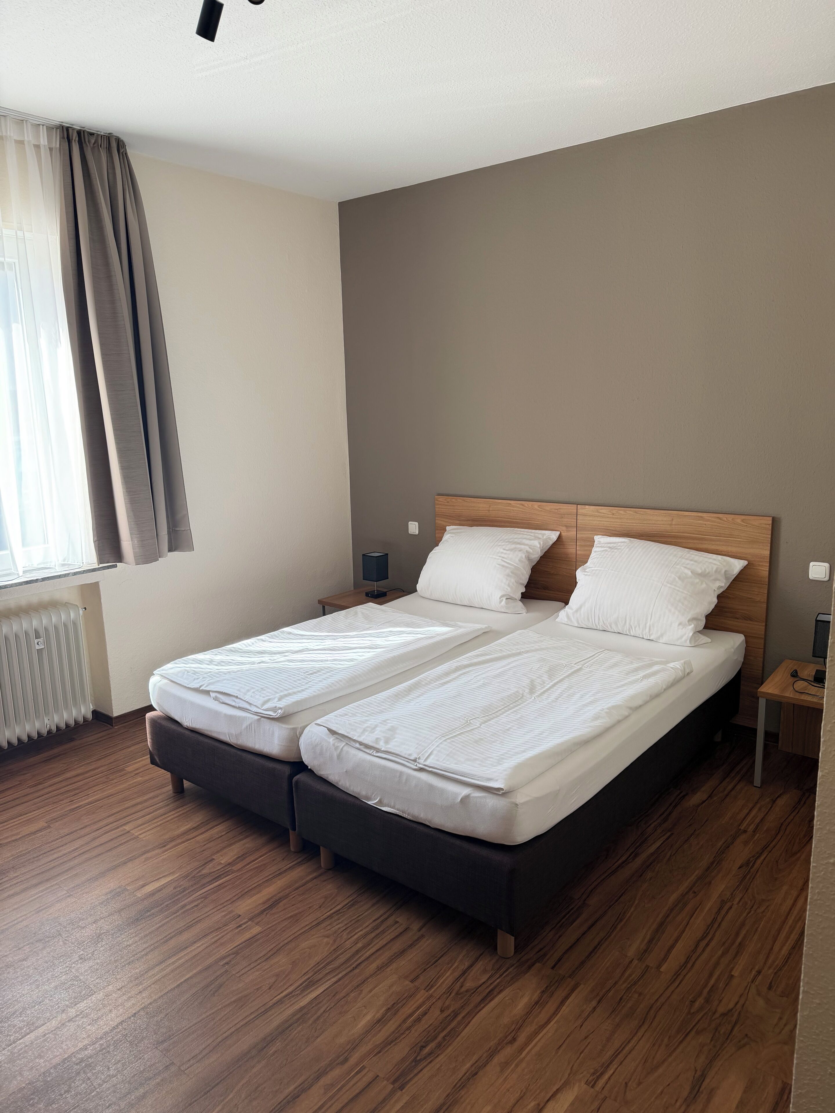 A double bed with a wooden headboard in a room with a radiator, a nightstand, and a window with curtains.