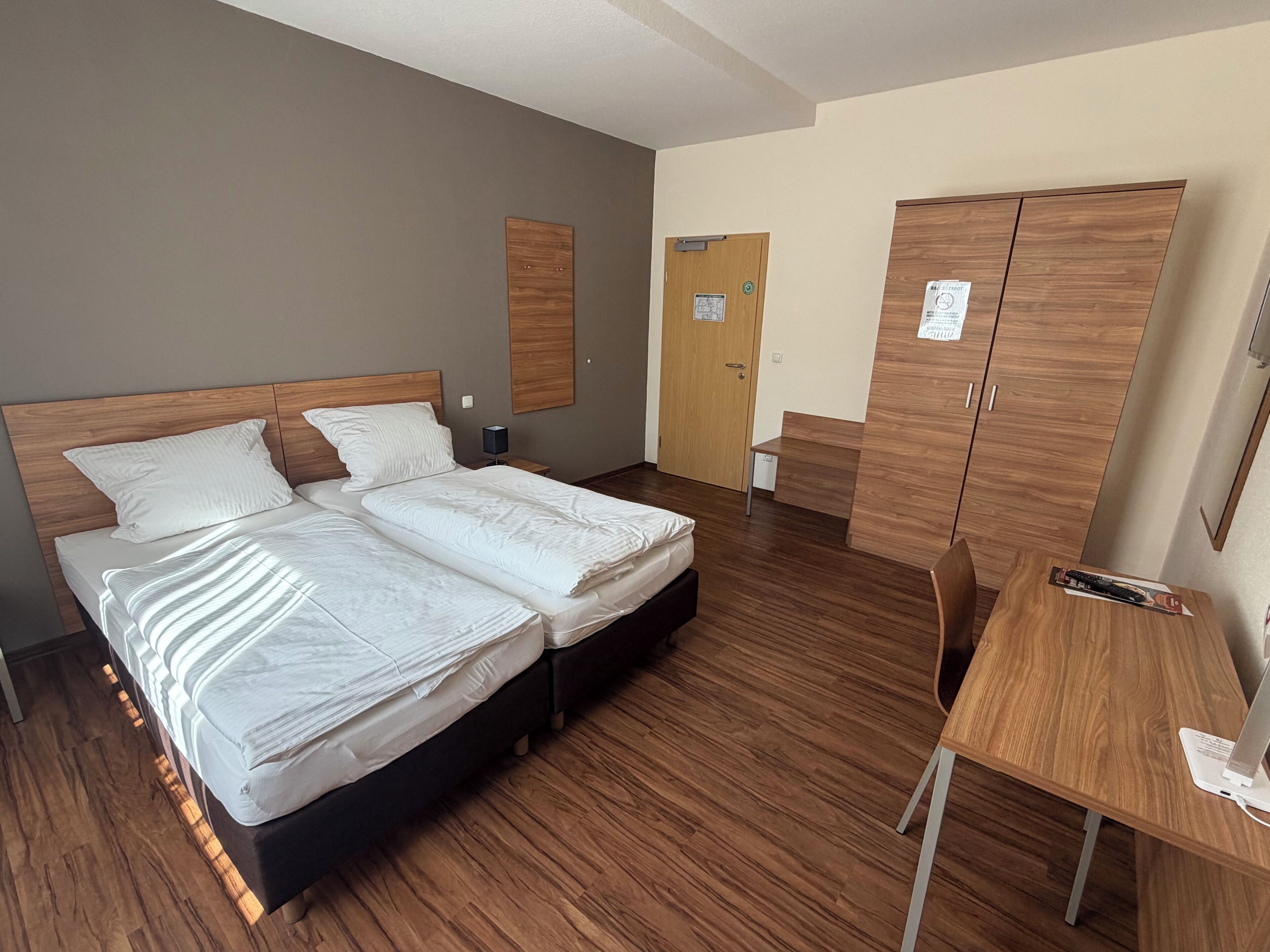 Habitación de hotel con cama doble, escritorio con computadora, armario y puerta con un cartel.