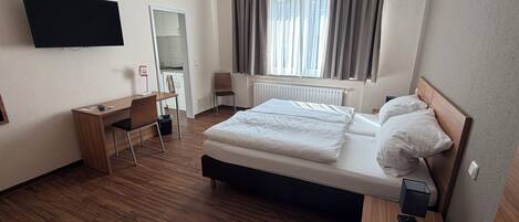 A hotel room with a bed, a desk, a chair, a TV mounted on the wall, and a window with curtains.