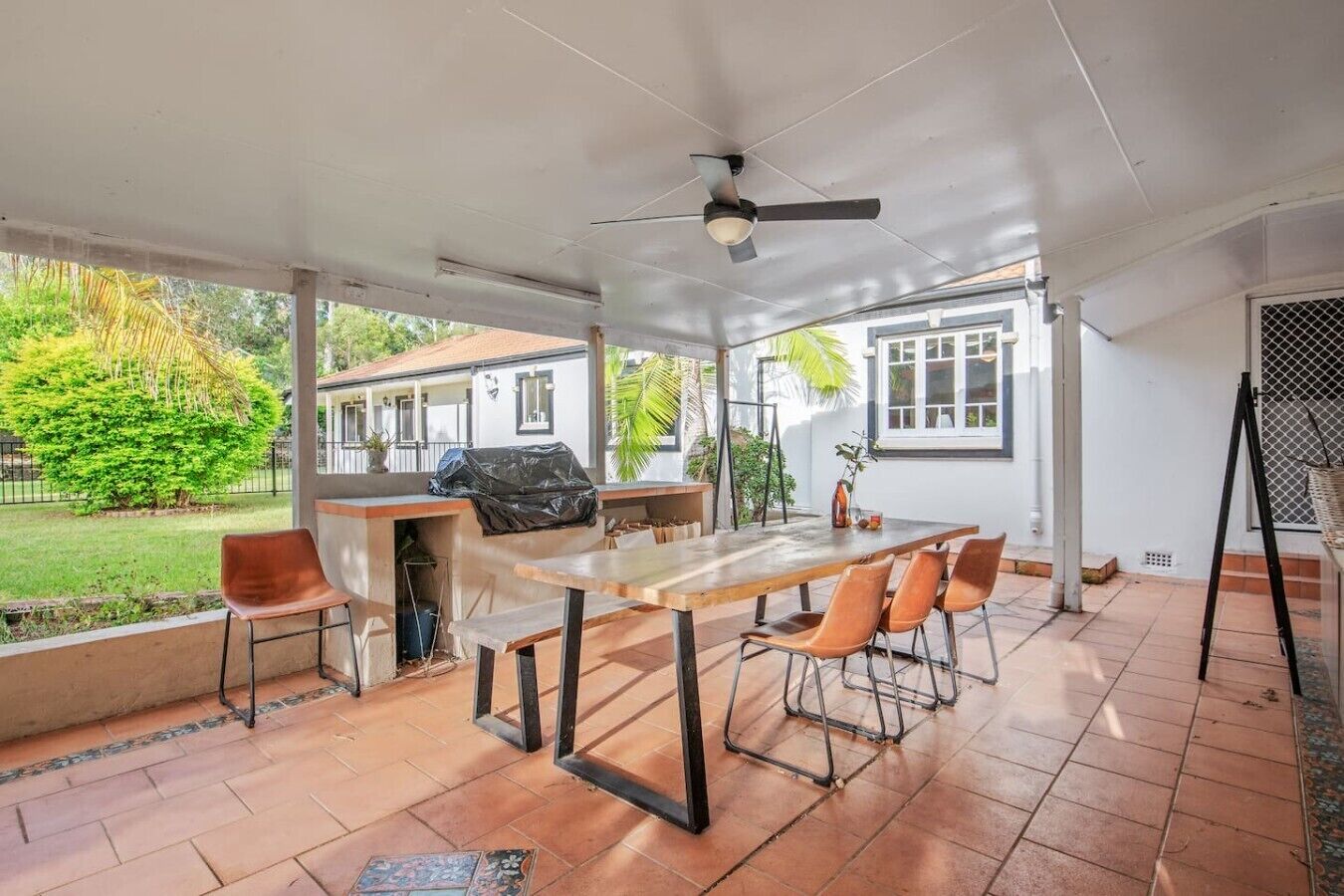 Patio esterno con tavolo e sedie, una ventola a soffitto e vista su una casa e vegetazione.