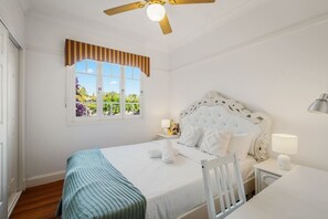 A bedroom with a bed, bedside lamps, a window with curtains, a ceiling fan, and a view of greenery outside.